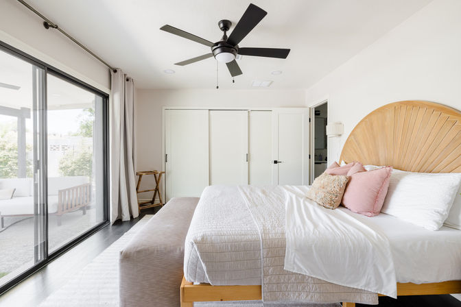 Bright modern bedroom with sliding glass doors to an outdoor patio, wooden sunburst headboard, white bedding with pink accent pillows, bench at foot and black ceiling fan