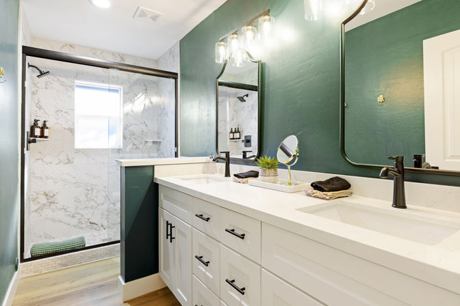 Sleek modern bathroom with white double-sink vanity, black faucets and hardware, deep green accent wall, round-edged mirrors, and glass-enclosed marble walk-in shower.