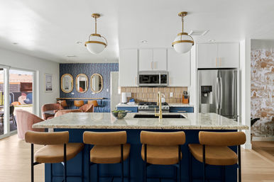 Sunlit open-concept modern kitchen with navy island and granite countertop, gold faucet and pendant lights, four tan leather bar stools, stainless steel fridge, blue wallpapered dining nook and sliding patio doors.