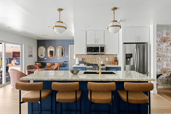 Sunlit open-concept modern kitchen with navy island and granite countertop, gold faucet and pendant lights, four tan leather bar stools, stainless steel fridge, blue wallpapered dining nook and sliding patio doors.