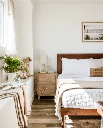 Sunlit beachy bedroom with a wooden bed, white quilt and beige throw, carved wood nightstand with lamp, potted fern by the window and framed ocean print on the wall.