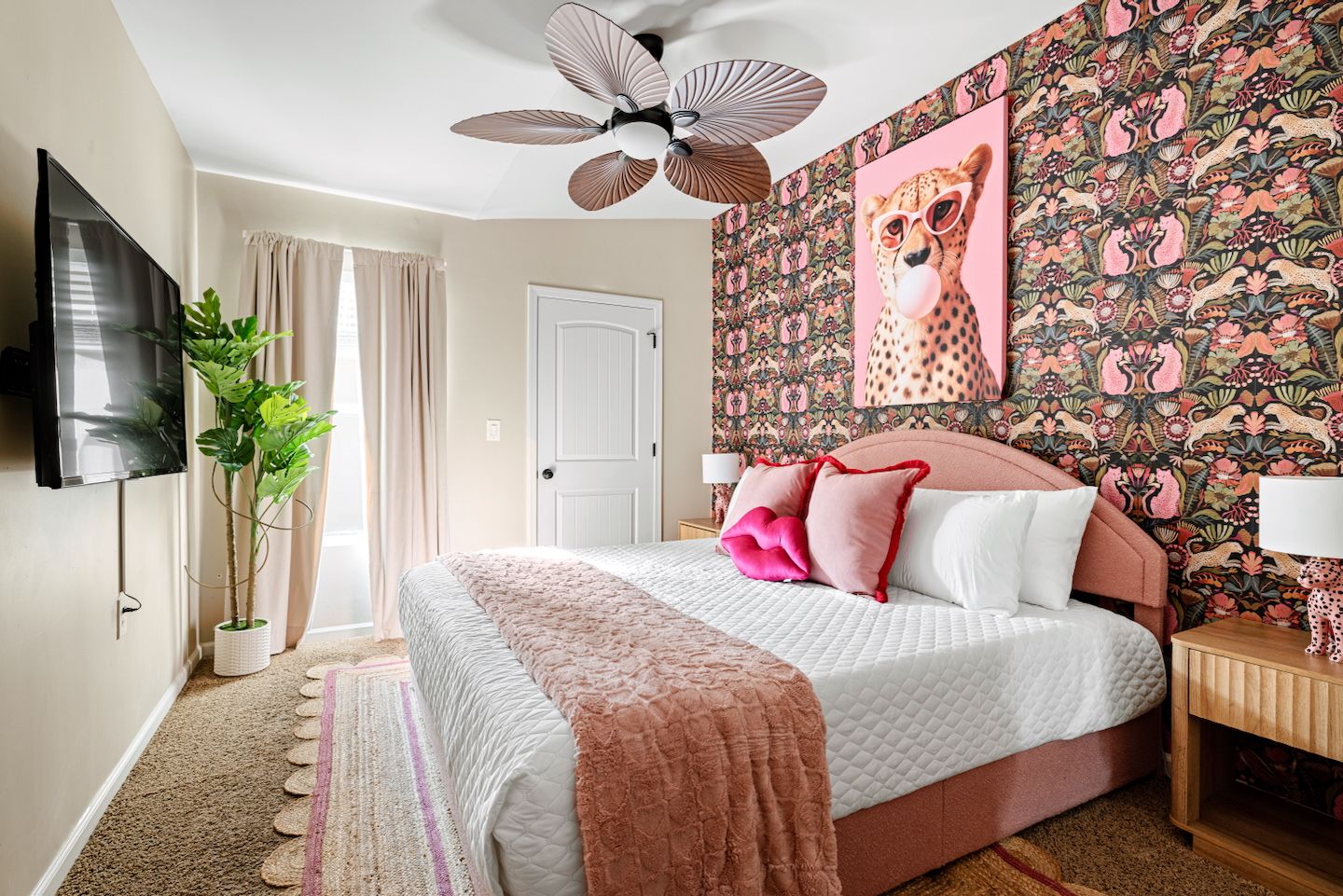Bright pink-accent bedroom with large upholstered bed, floral-animal patterned wallpaper and cheetah pop-art, plush white bedding with pink throw and pillows, leafy potted plant, wooden nightstands and decorative ceiling fan.