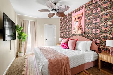 Bright pink-accent bedroom with large upholstered bed, floral-animal patterned wallpaper and cheetah pop-art, plush white bedding with pink throw and pillows, leafy potted plant, wooden nightstands and decorative ceiling fan.