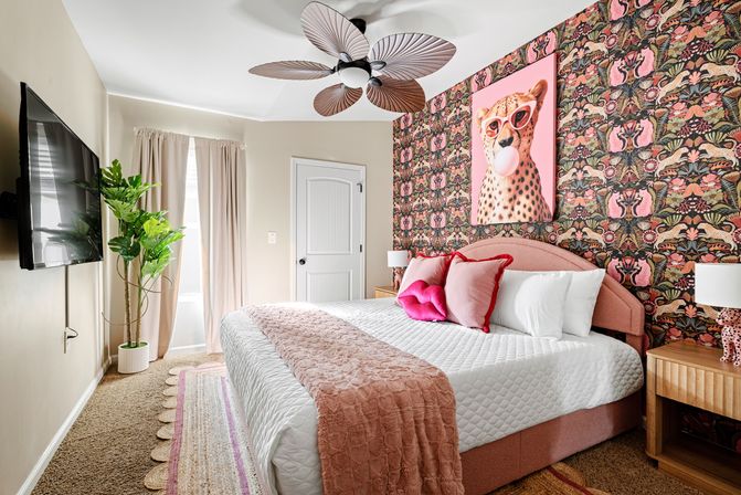 Bright pink-accent bedroom with large upholstered bed, floral-animal patterned wallpaper and cheetah pop-art, plush white bedding with pink throw and pillows, leafy potted plant, wooden nightstands and decorative ceiling fan.