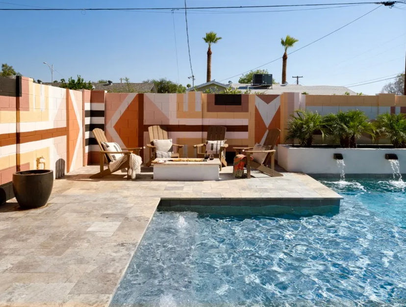 Sunny backyard pool patio with sparkling blue water, tiled deck, Adirondack chairs around a low fire pit, geometric painted cinderblock wall and palm trees