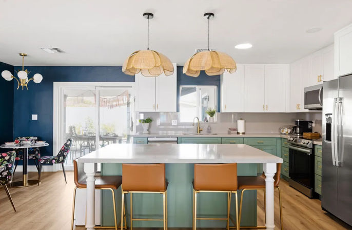 Bright open-concept modern kitchen with white island, teal lower cabinets, tan leather bar stools, rattan pendant lights, stainless appliances and navy dining nook