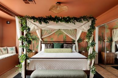 Lush tropical-inspired bedroom with a four-poster canopy bed draped in sheer curtains and greenery, palm mural on terracotta walls, patterned pillows, window seat, mirrored closet and textured bench.