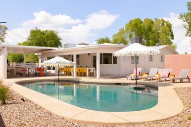 Sunny suburban backyard with a kidney-shaped pool, scalloped-edge umbrellas in the water, lounge chairs with pink towels, yellow dining chairs under a covered patio, and gravel landscaping.