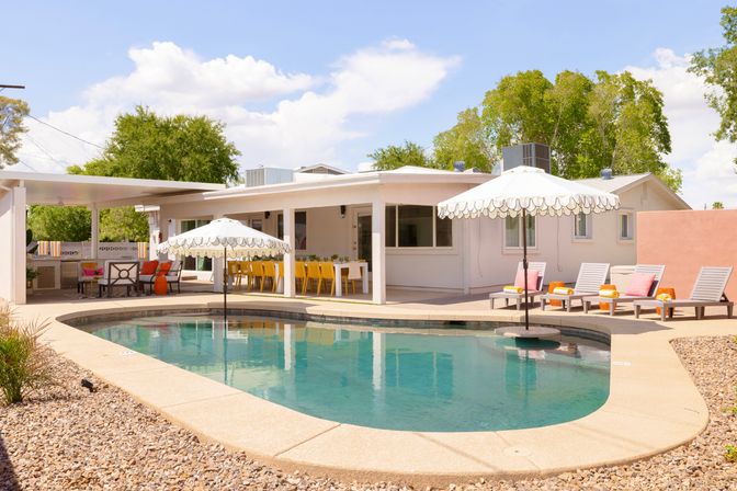 Sunny suburban backyard with a kidney-shaped pool, scalloped-edge umbrellas in the water, lounge chairs with pink towels, yellow dining chairs under a covered patio, and gravel landscaping.
