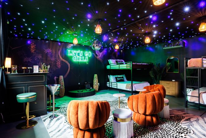 Vibrant indoor party suite with neon "LET'S GO GIRLS" sign, starry ceiling lights and disco ball, orange velvet lounge chairs and bunk beds