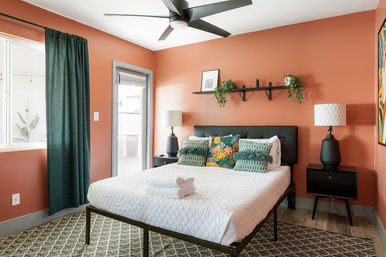 Bright modern bedroom with terracotta walls, black upholstered headboard, white quilted bed topped with folded towels and tropical green accent pillows, dark nightstands with matching lamps, patterned rug, ceiling fan, and green curtains by a sliding patio door.
