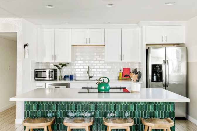 Bright modern white kitchen with glossy subway tile backsplash, green arched-tile island and white countertop, four wooden stools, stainless steel refrigerator and microwave, and a green kettle on the island cooktop.