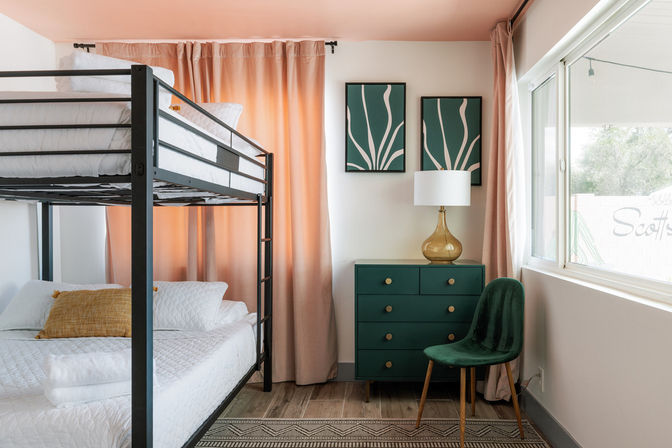 Cheerful sunlit bedroom with black metal bunk bed, teal dresser with gold lamp, green velvet chair and coral curtains