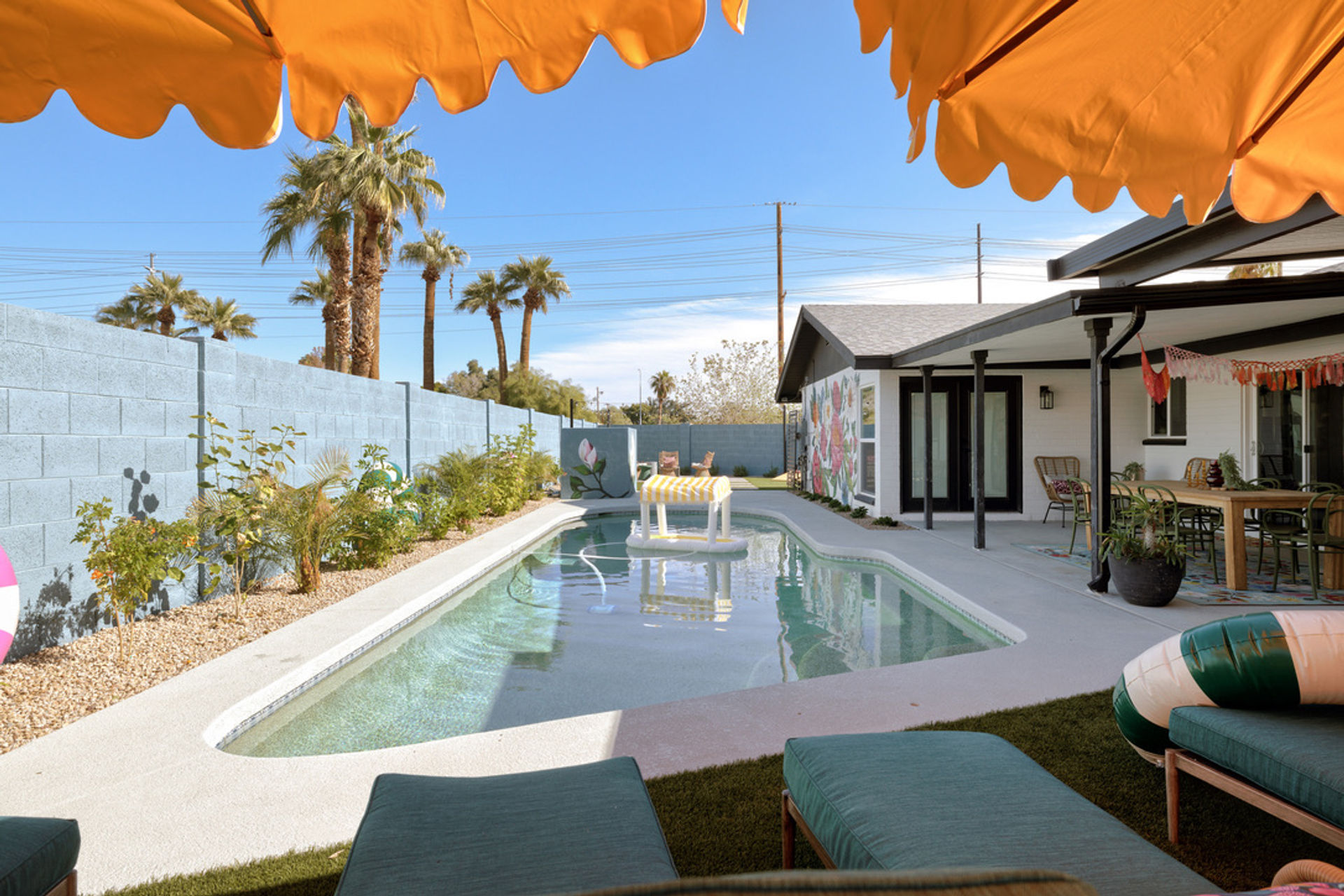 Sunny Southwestern backyard pool with palm trees, orange umbrella shade, floating lounger, covered patio dining area and lounge seating beside a blue block privacy wall.