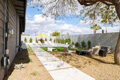 Sunny modern backyard with a rectangular pool, white lounge chairs and yellow umbrella, concrete pavers across artificial turf, outdoor seating under a shade tree, palm trees and a gray block privacy wall.