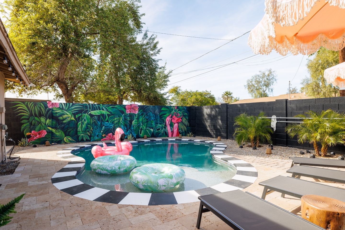 Sunny suburban backyard pool with black-and-white tiled edge, vibrant tropical mural with oversized leaves and pink flowers, a pink inflatable flamingo and green leaf floats, lounge chairs and fringed umbrellas