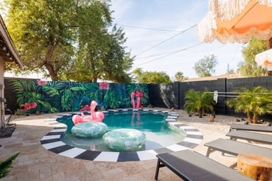 Sunny suburban backyard pool with black-and-white tiled edge, vibrant tropical mural with oversized leaves and pink flowers, a pink inflatable flamingo and green leaf floats, lounge chairs and fringed umbrellas
