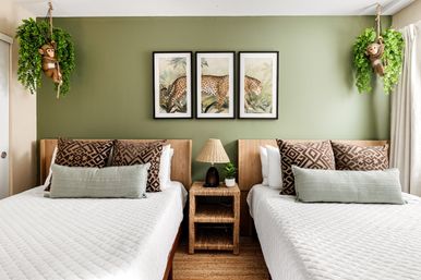 Twin bedroom with sage green accent wall, three-panel leopard artwork, rattan nightstand and lamp, patterned pillows and playful hanging monkey planters.