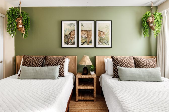 Twin bedroom with sage green accent wall, three-panel leopard artwork, rattan nightstand and lamp, patterned pillows and playful hanging monkey planters.