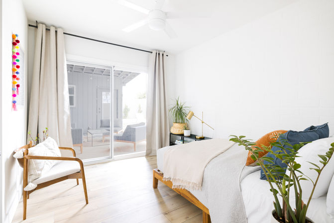 Bright, minimalist bedroom with mid-century wooden bed and chair, neutral linens and colorful throw pillows, potted plants, a gold bedside lamp, and large sliding glass doors opening to a sunlit patio.