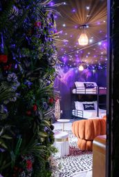 Whimsical boho-chic lounge with bunk beds, plush orange pouf, leopard-print rug, small round tables, lush vertical garden and purple twinkling star ceiling lights.