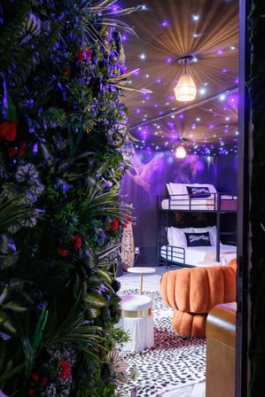 Whimsical boho-chic lounge with bunk beds, plush orange pouf, leopard-print rug, small round tables, lush vertical garden and purple twinkling star ceiling lights.