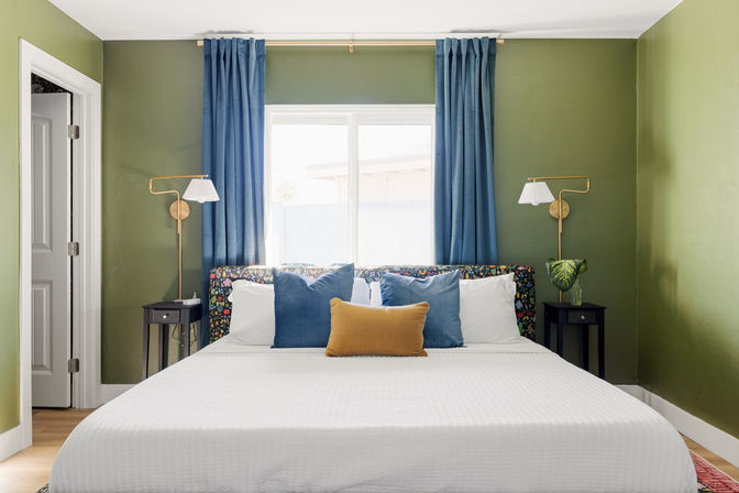 Cozy bedroom with olive-green walls, blue curtains framing a sunlit window, floral headboard, white king bed with blue and mustard accent pillows and brass wall sconces