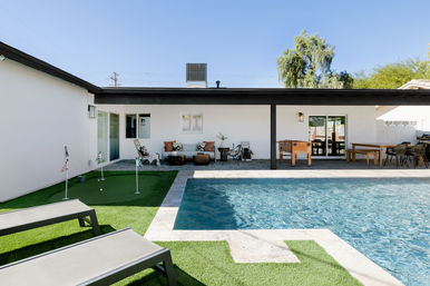 Sunny backyard with rectangular pool, small putting green and gray lounge chairs, covered patio featuring cushioned sofa seating, foosball table and outdoor dining set