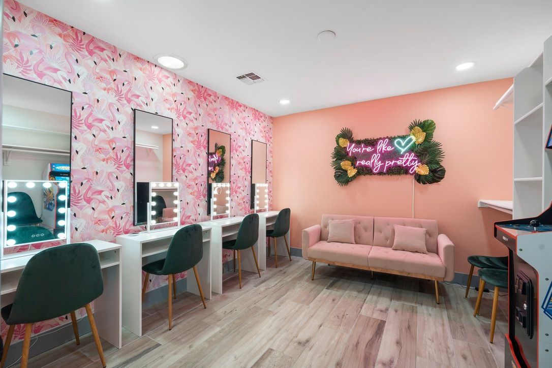 Bright pink boutique makeup studio interior with flamingo wallpaper, three lighted vanity mirrors and green velvet chairs, blush tufted sofa, neon sign reading "You're like really pretty," tropical foliage accents and wood-look flooring.