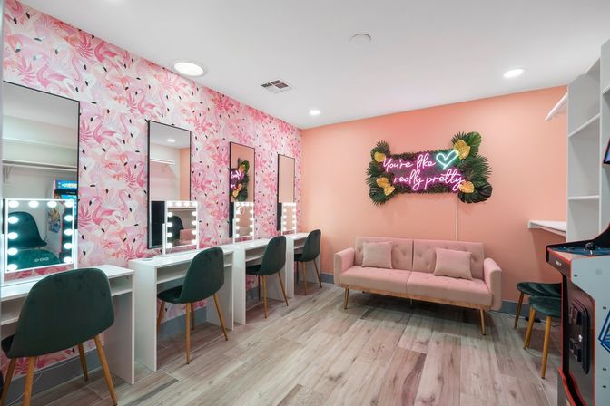 Bright pink boutique makeup studio interior with flamingo wallpaper, three lighted vanity mirrors and green velvet chairs, blush tufted sofa, neon sign reading "You're like really pretty," tropical foliage accents and wood-look flooring.