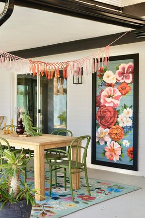 Bright covered patio dining area with wooden table, green chairs, colorful floral rug, large framed floral mural, potted plants and pink tassel garland overhead.