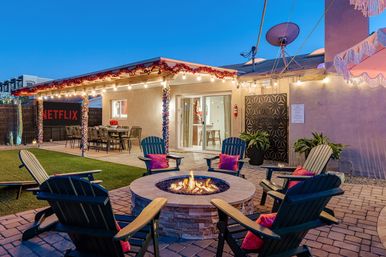 Cozy suburban backyard patio at dusk with a circular stone fire pit surrounded by Adirondack chairs and bright pillows, string lights and garland over a covered dining area with an outdoor movie screen, potted plants, and sliding glass doors.