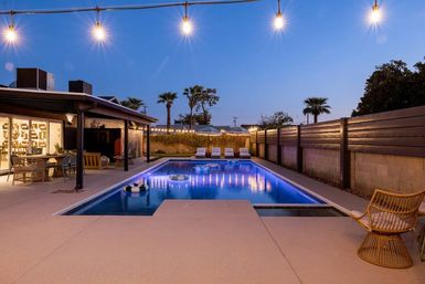 Resort-style backyard at dusk with a glowing rectangular swimming pool and floating rings, string lights overhead, covered patio with dining and lounge seating, poolside sunbeds and palm trees in the background.