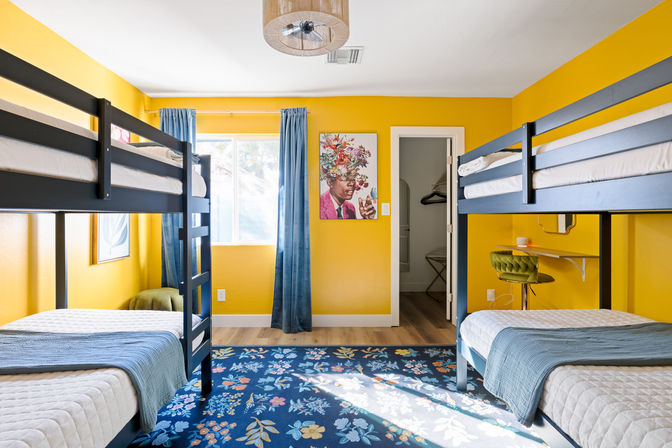 Sunlit family bunk room in a vacation rental with bright yellow walls, two navy bunk beds with blue quilts, floral blue area rug, blue curtains, a green swivel desk chair and colorful floral portrait