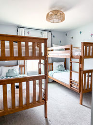 Bright kids' bedroom with two wooden bunk beds, light-gray walls with hot-air-balloon decals, cloud-shaped pillows, white curtains, woven ceiling light, and neutral carpet