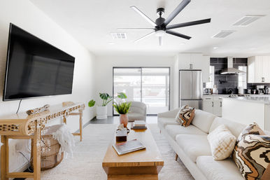 Sunlit modern open-concept living room and kitchen with a neutral sofa, patterned throw pillows, wooden coffee table, wall-mounted flat-screen TV, stainless-steel refrigerator, sliding glass patio doors, large black ceiling fan, and potted plants.