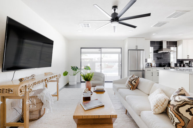Sunlit modern open-concept living room and kitchen with a neutral sofa, patterned throw pillows, wooden coffee table, wall-mounted flat-screen TV, stainless-steel refrigerator, sliding glass patio doors, large black ceiling fan, and potted plants.