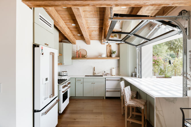 Bright open-concept kitchen with exposed wood beams, mint-green cabinets, marble countertops and a garage-style glass door folding open to a sunlit garden, plus a breakfast bar with woven stools.