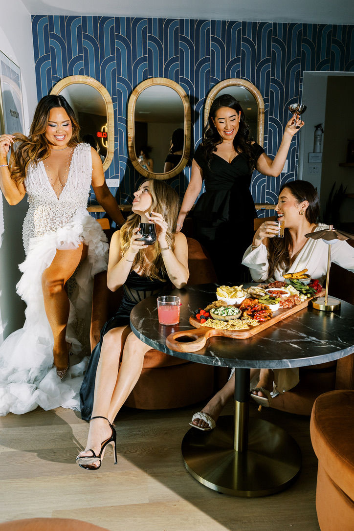 Four dressed-up women laughing and toasting with cocktails around a marble-topped table featuring a charcuterie board in a stylish indoor lounge with navy Art Deco wallpaper and oval mirrors.