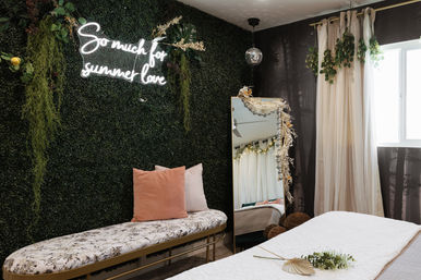 Boho bedroom nook with a faux boxwood plant wall and neon sign reading "So much for summer love", floral-upholstered bench with blush pillows, full-length mirror draped in dried flowers, sheer curtains by a bright window and a small disco ball.