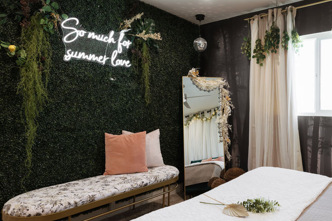 Boho bedroom nook with a faux boxwood plant wall and neon sign reading "So much for summer love", floral-upholstered bench with blush pillows, full-length mirror draped in dried flowers, sheer curtains by a bright window and a small disco ball.