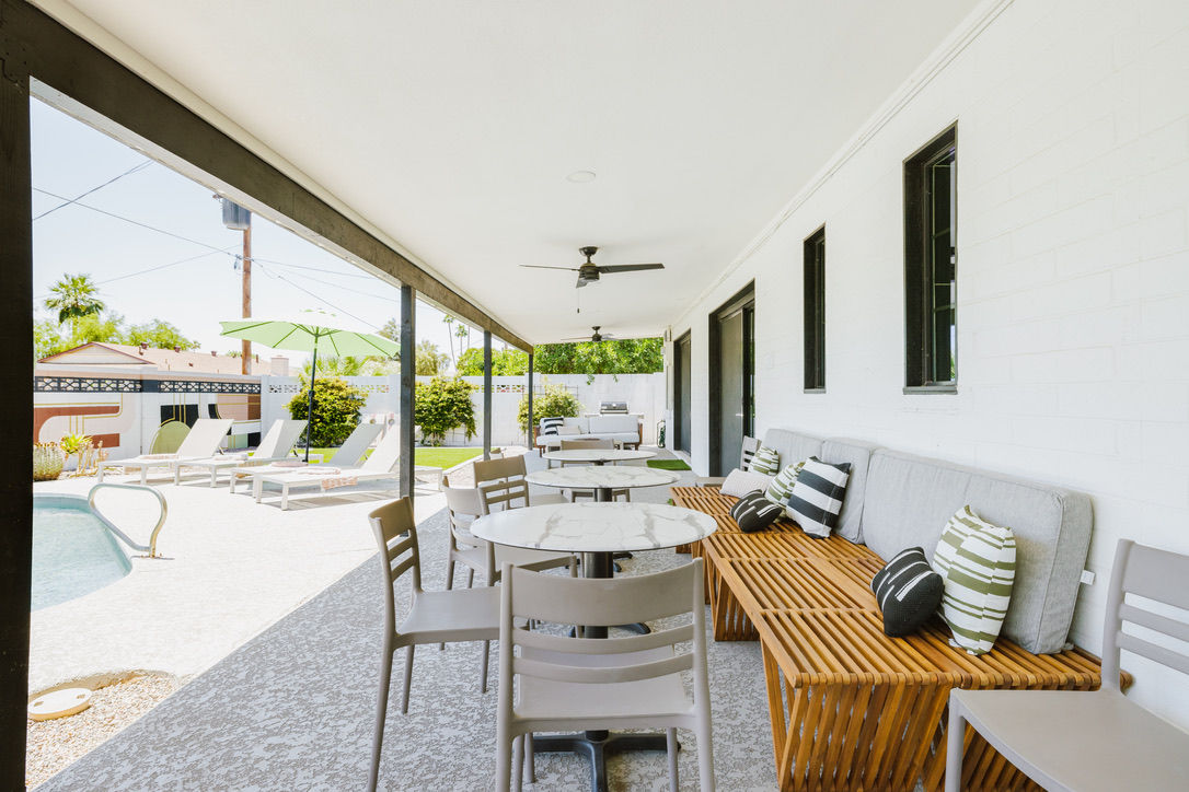 Covered poolside suburban backyard patio with round marble-top tables, wooden slatted bench with striped throw pillows, ceiling fans overhead and lounge chairs under a green umbrella on a sunny day