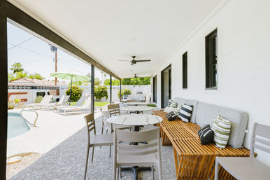 Covered poolside suburban backyard patio with round marble-top tables, wooden slatted bench with striped throw pillows, ceiling fans overhead and lounge chairs under a green umbrella on a sunny day