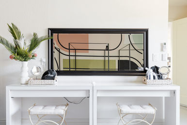 Bright modern dual white vanity with tufted stools, large black-framed abstract mirror, woven trays with hair tools, and a tall tropical floral arrangement