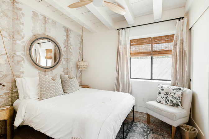 Bright cozy boho bedroom with white-beamed ceiling, queen bed in white linens and patterned pillows, round wall mirror over textured wallpaper, hanging macramé pendant, bamboo blinds and a plush accent chair