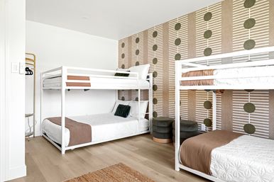 Bright modern shared bedroom with two white metal bunk beds, quilted white bedding and brown throws, light wood floors, geometric wallpaper with green dots, velvet round ottomans and an open gold clothing rack.