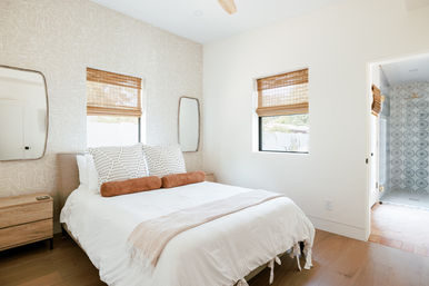 Sunlit modern bedroom with white linens, textured accent wall, wood floors and bamboo window shades, rust bolster pillows and a wood nightstand, with a glimpse of a patterned-tile ensuite bathroom.