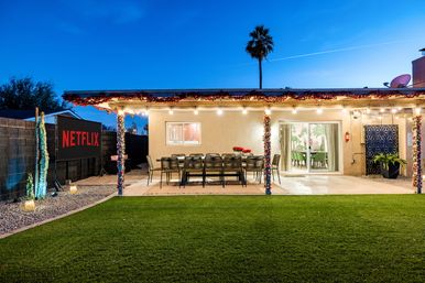Dusk backyard patio ready for movie night with string lights and garlands, long outdoor dining table, sliding glass doors, palm tree, green lawn and an outdoor screen showing a streaming logo.