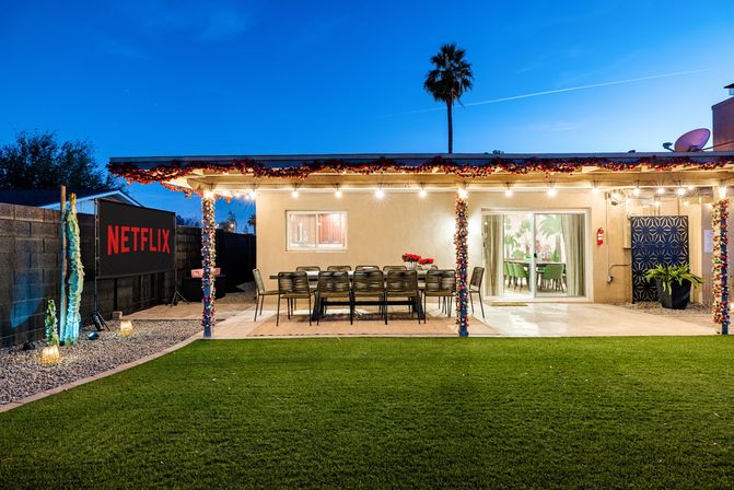 Dusk backyard patio ready for movie night with string lights and garlands, long outdoor dining table, sliding glass doors, palm tree, green lawn and an outdoor screen showing a streaming logo.
