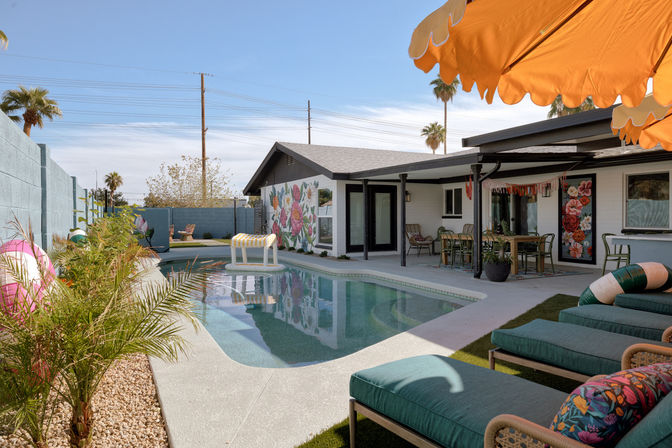 Sunlit backyard pool oasis with teal lounge chairs, orange umbrella, floral mural on the house, covered dining patio, palm trees and a striped pool float